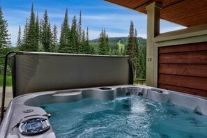 Un jacuzzi con panel de control, rodeado por una terraza de madera y vistas a altos árboles y una montaña.