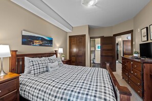 Un dormitorio con una cama grande, cabecera de madera, una mesita de noche con lámpara, cómoda y un cuadro de un paisaje montañoso en la pared.