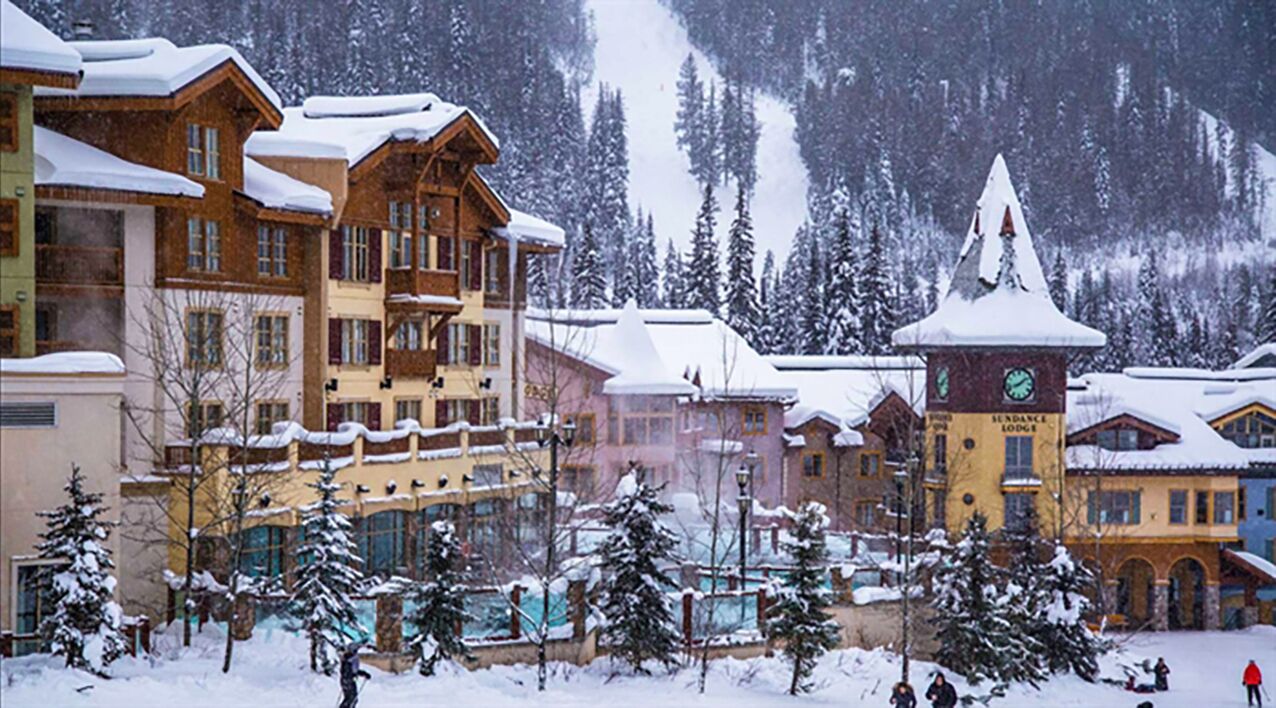 Une station de ski enneigée, avec des bâtiments en bois, une tour imposante qui fait office de refuge, et une piste de ski en arrière-plan.