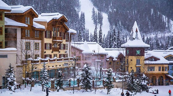 Un resort cubierto de nieve con edificios de madera, una imponente torre de albergue y una pista de esquí al fondo.
