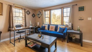 A living room with a blue sofa, a wooden coffee table, and a desk with a chair. There are two windows with curtains, a wall clock, and a welcome sign. - Luxury Home Central to Farmington, Plainville & Downtown New Britain (New Britain)