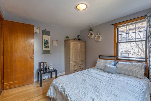 A bedroom with a wooden door, a bed with white bedding, a small table with a vase, and a window with a view of bare trees.