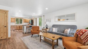 Ein modernes Wohnzimmer mit Sofa, Couchtisch und Sesseln, im Hintergrund eine Küche und ein großes Fenster.