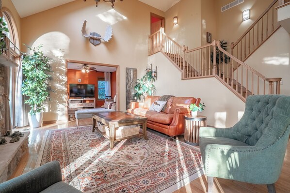 A spacious living room with a large wooden coffee table, a leather sofa, and a green armchair. A staircase with wooden railings leads to an upper level.