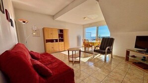 Een woonkamer met een rode bank, een eettafel met stoelen, een tv-meubel en een groot raam met uitzicht op het buitengebied.