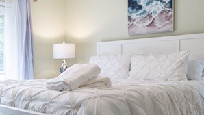 A neatly made bed with a white duvet and pillows, a bedside lamp, and a painting of a wave on the wall. - Family Entertainment Beach House, Hot tub, Nature Park, Fire pit, Pizza Oven (Emerald Isle)