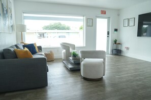 A modern hotel lobby with a grey sofa, a white armchair, and a small table with a plant. There is a large window providing a view of the outside, and an exit sign is visible on the wall.
