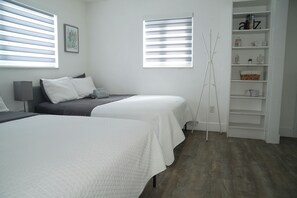 A hotel room with two beds, a bedside table, a lamp, a bookshelf, and a window with blinds.