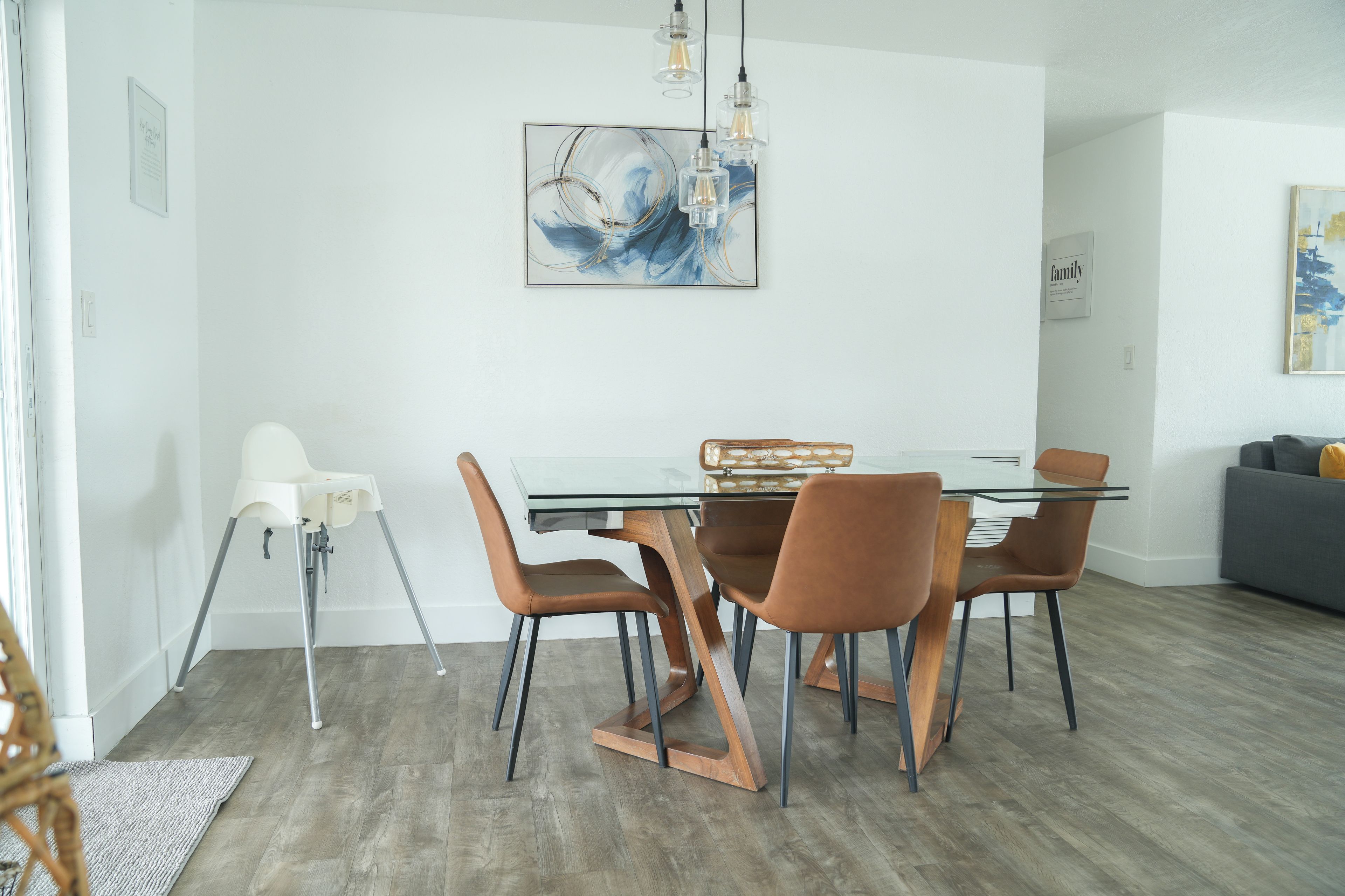 A modern dining area with a glass table, brown chairs, and a painting above.