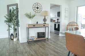 A modern living room with a wooden floor, a console table with a lamp, a wall clock, and a framed quote.