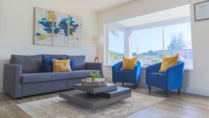 A living room with a grey sofa, blue armchairs, a coffee table, and a large window offering a view of the outside.