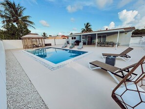 Piscina retangular com um deck branco, rodeada por espreguiçadeiras e uma área de estar coberta.
