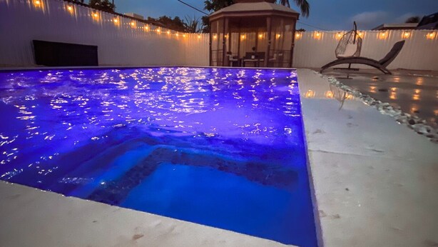 An outdoor swimming pool with blue lighting, surrounded by a white wall and palm trees.