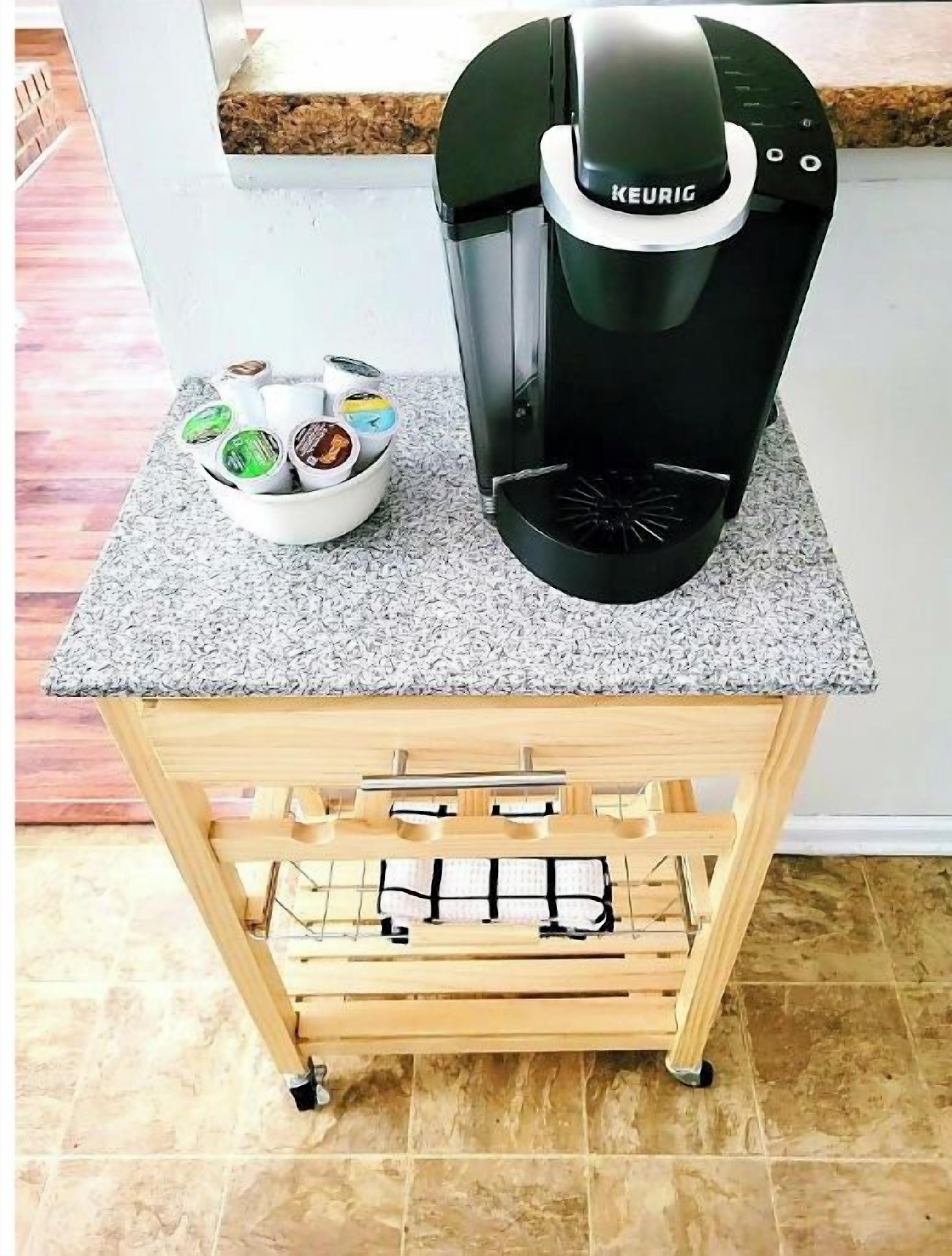 Une machine à café Keurig posée sur un comptoir en granit, avec un porte-capsules à café, et un chariot en bois muni d’une étagère métallique en dessous.