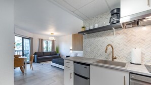 A modern kitchen with a dishwasher, a coffee maker, and a sink. There is a dining area with a table and chairs, and a living room with a sofa and a lamp. - NEW - Spacious Studio close to Theme Parks (Orlando)