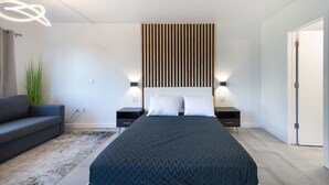 A modern bedroom with a large bed, a dark-colored headboard, two bedside tables with lamps, a grey sofa, and a tall plant. - NEW - Spacious Studio close to Theme Parks (Orlando)