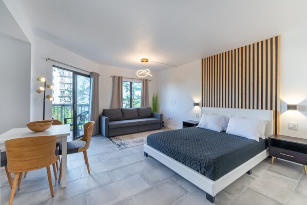 A modern bedroom with a large bed, a desk with chairs, a sofa, and a balcony with a view. - NEW - Spacious Studio close to Theme Parks (Orlando)