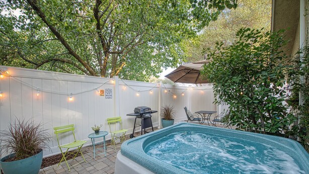 A hot tub in a backyard with a white fence, string lights, and outdoor furniture.