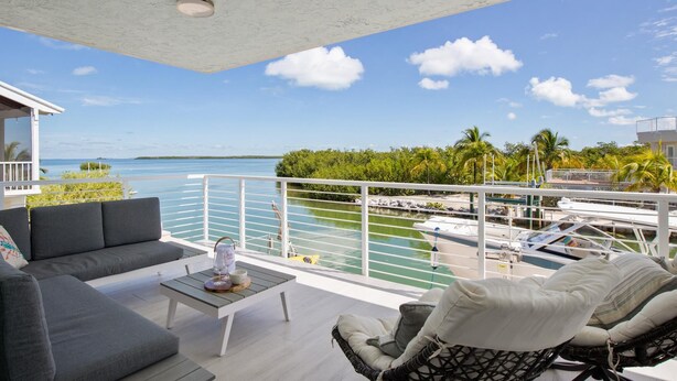Een balkon met een bank, stoel en tafel, met uitzicht op een jachthaven vol boten en een helderblauwe lucht.
