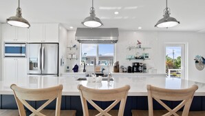 Een moderne keuken met witte kasten, roestvrijstalen apparatuur, een groot eiland met barstoelen, hanglampen en uitzicht naar buiten.
