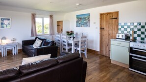 A living room with a brown leather sofa, a dining area with white chairs, a kitchen with a stove, and a window with curtains. - Harbour Lodge (Port Isaac)