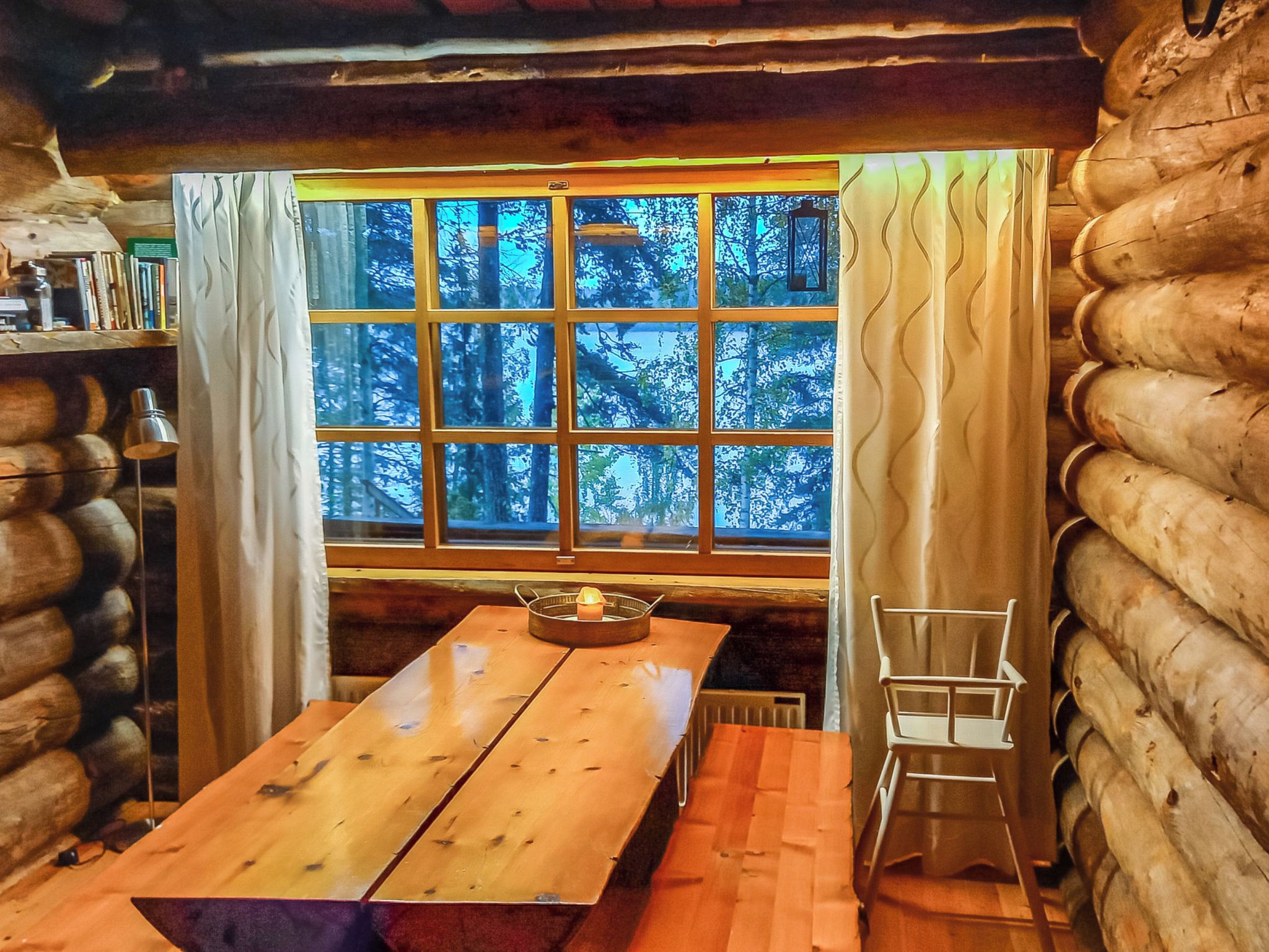 A log cabin interior with a wooden table, a chair, and a window with a view of trees.