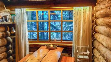 A log cabin interior with a wooden table, a chair, and a window with a view of trees.
