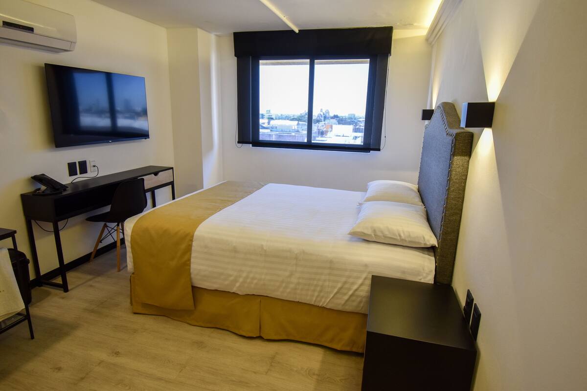 Una habitación de hotel con cama, escritorio, silla, televisión y ventana con vista a la ciudad.