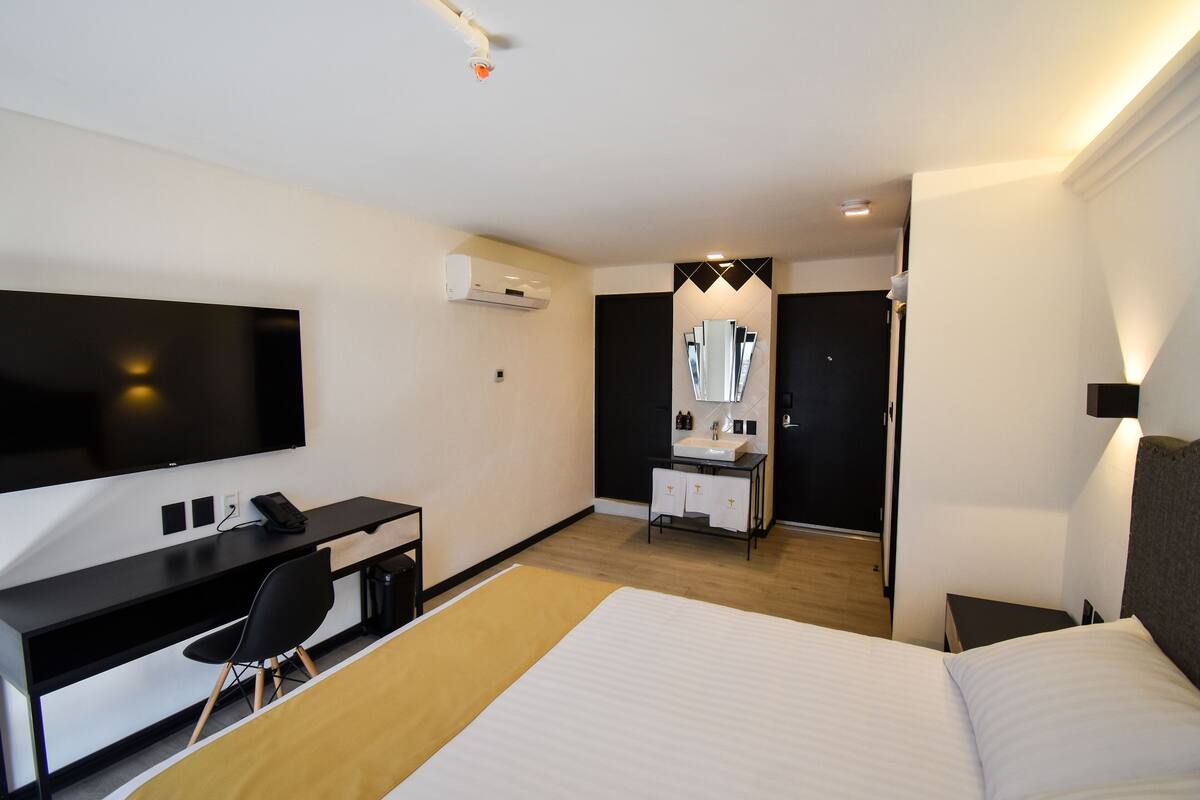 Una habitación de hotel moderna con una cama, un escritorio con una silla, un televisor, un baño con espejo y una mesita de noche.