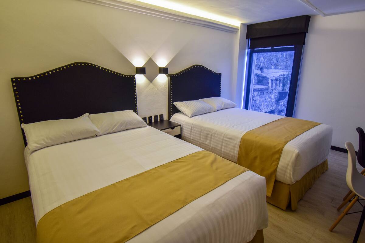 Habitación de hotel con dos camas, cada una con cabecero, ropa de cama blanca y un detalle amarillo. Se aprecia una ventana con vista a un edificio y una silla en la esquina.