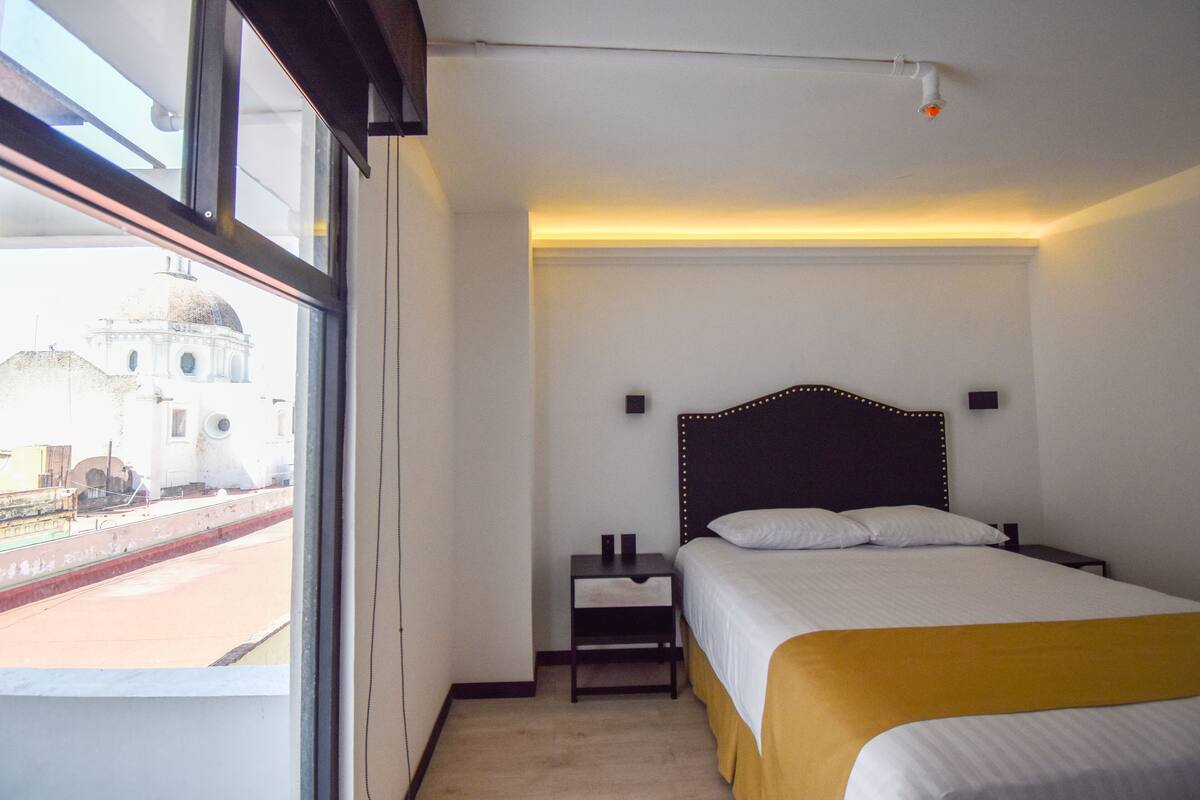 Una habitación de hotel con un ventanal grande, una cama con cabecera, una mesita de noche y una lámpara fijada a la pared.