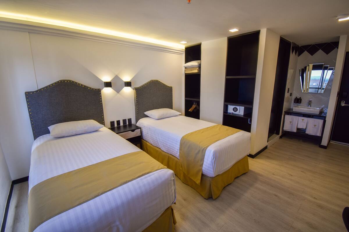 Habitación de hotel con dos camas, una mesita de noche, armario empotrado, tocador con espejo y un pequeño rincón de descanso.
