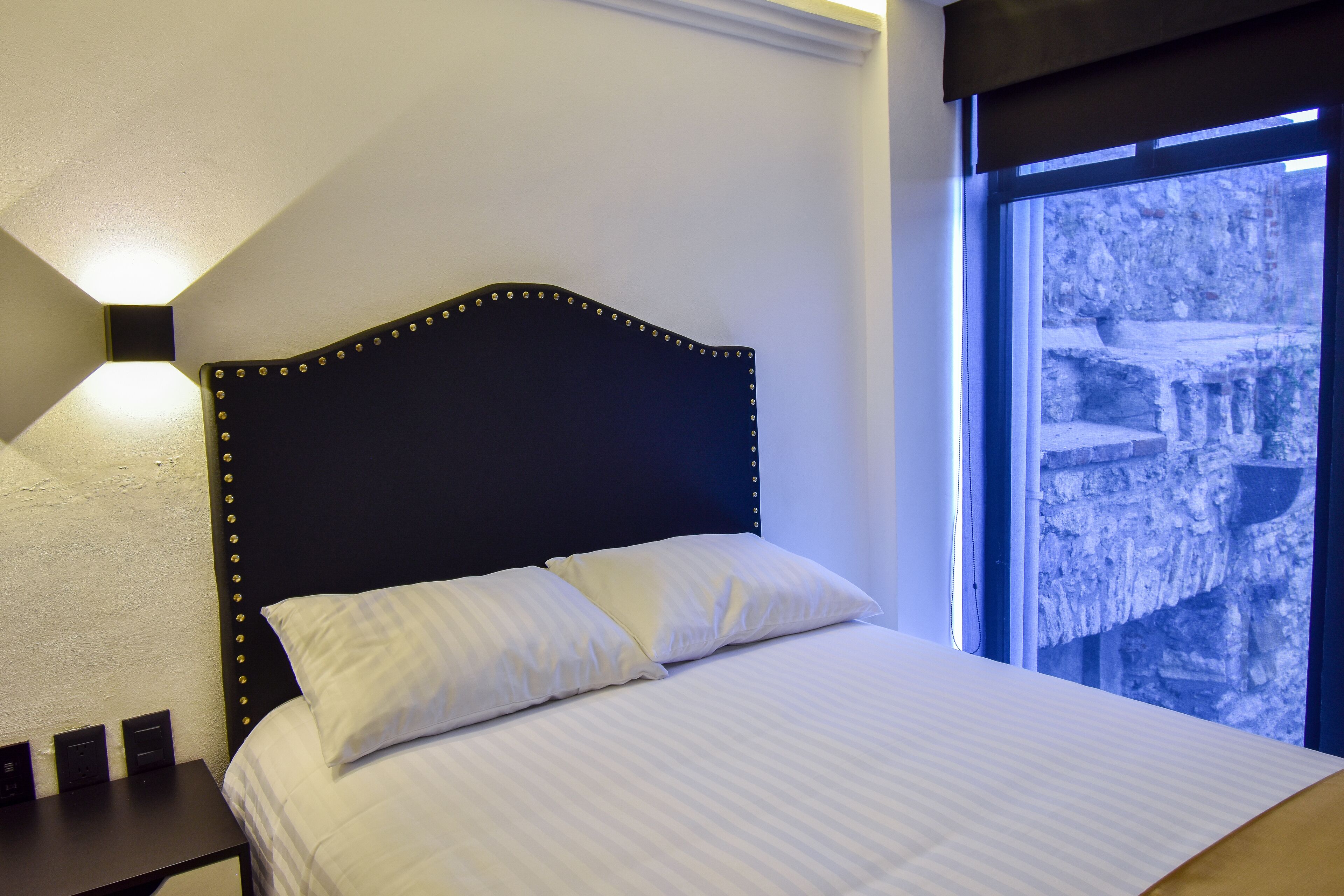 Una cama bien hecha con sábanas blancas y dos almohadas, un cabecero oscuro con tachuelas decorativas, una lámpara de noche y una ventana con vistas a una estructura de piedra exterior.