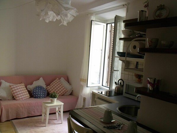 A cozy living room with a pink sofa, a small white table with a plant, and a kitchen area with a sink and stove.