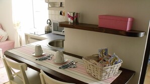 Eine kleine Küche mit einem Holztisch, weißen Stühlen, einem Edelstahl-Wasserkocher, einem Korb voller Lebensmittel und einer rosa Kiste auf einem Regal.
