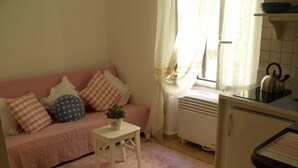 Ein gemütliches Wohnzimmer mit einem rosafarbenen Sofa, einem kleinen weißen Tisch mit einer Pflanze und einer Küchenzeile mit einem Wasserkocher.