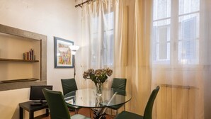 A dining area with a round glass table, four green chairs, a vase with flowers, a bookshelf, a framed picture, and a window with sheer curtains. - Pitti Studio apartment by Mmega (Firenze)