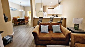 Un salon chaleureux avec un canapé marron, un coin repas avec des chaises en osier et une cuisine avec des meubles blancs.