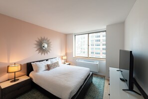Ein Hotelzimmer mit Bett, Nachttischen, Lampe, Fernseher und Fenster mit Blick auf die Stadt.
