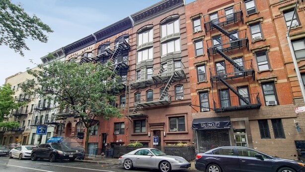A multi-story building with fire escapes, parked cars, and a tree in front.