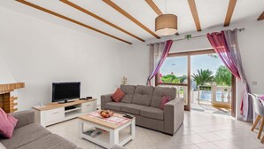 A modern living room with a sofa, a coffee table with fruit, a TV, and a balcony with curtains.