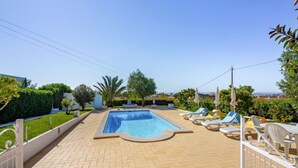 A rectangular swimming pool with a tiled deck, surrounded by lounge chairs and umbrellas, with a view of a distant landscape.