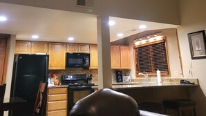 A spacious living area with a kitchen in the background, featuring wooden cabinets, a black refrigerator, and a microwave.