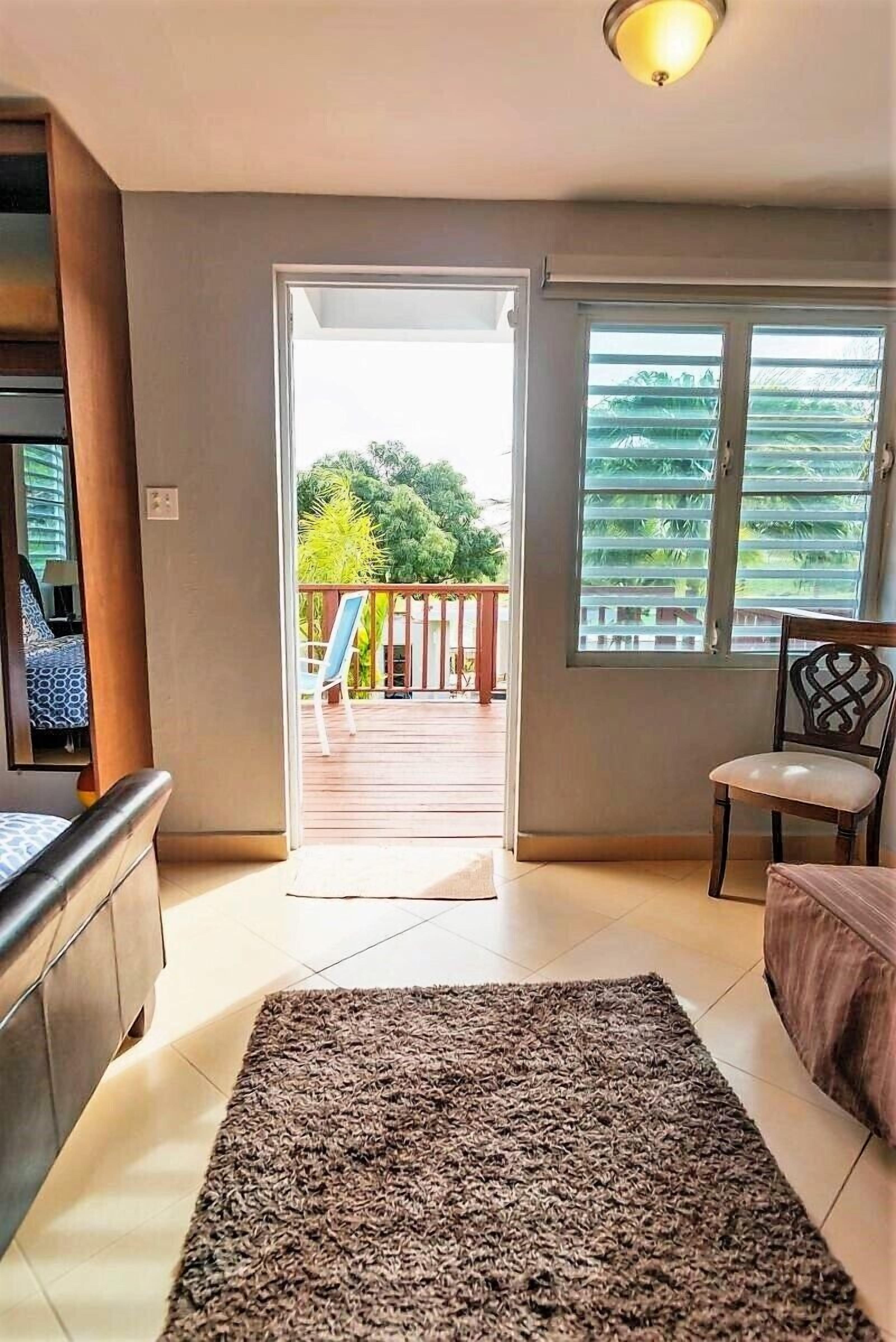 Ein Wohnzimmer mit einer Couch, einem Couchtisch und einem Teppich. Eine Tür führt zu einem Balkon mit Blick auf Grünflächen.