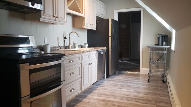 A kitchen with a stove, oven, dishwasher, and a refrigerator. There is a wooden floor and a metal cart. - Rosé All Day King Studio | Retro Vibes | Downtown| (Fredericton)