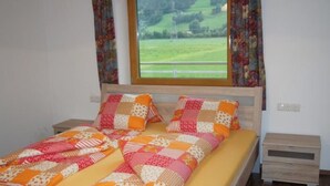 A bed with a quilt, a wooden headboard, a window with a view of a green landscape, and a nightstand with a drawer. - Apartment "Gertis Top 2" with Mountain View, Wi-Fi & Balcony (Stumm)