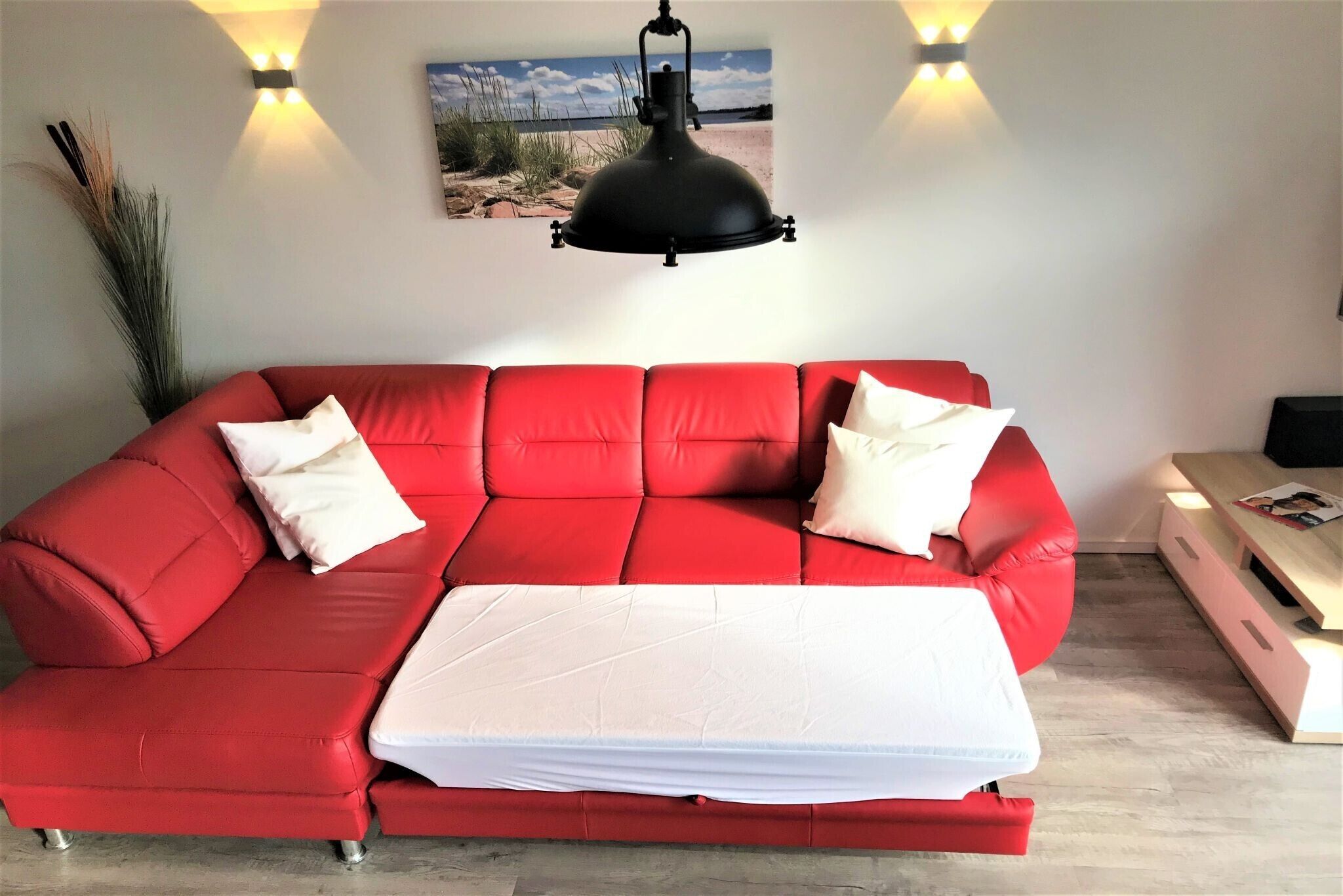 Un canapé rouge avec un matelas blanc, deux lampes fixées au mur et un tableau représentant une vue sur la plage.