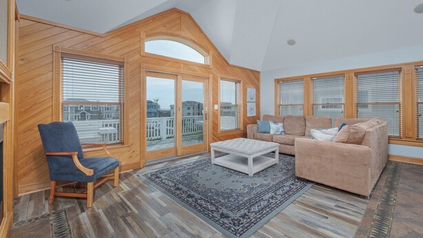 A spacious living room with wooden paneling, a large window with blinds, a blue armchair, a beige sofa, and a patterned rug. - 5566- Pearl Jam - Nags Head Retreat w/ Pool, Hot Tub & Theater (Nags Head)