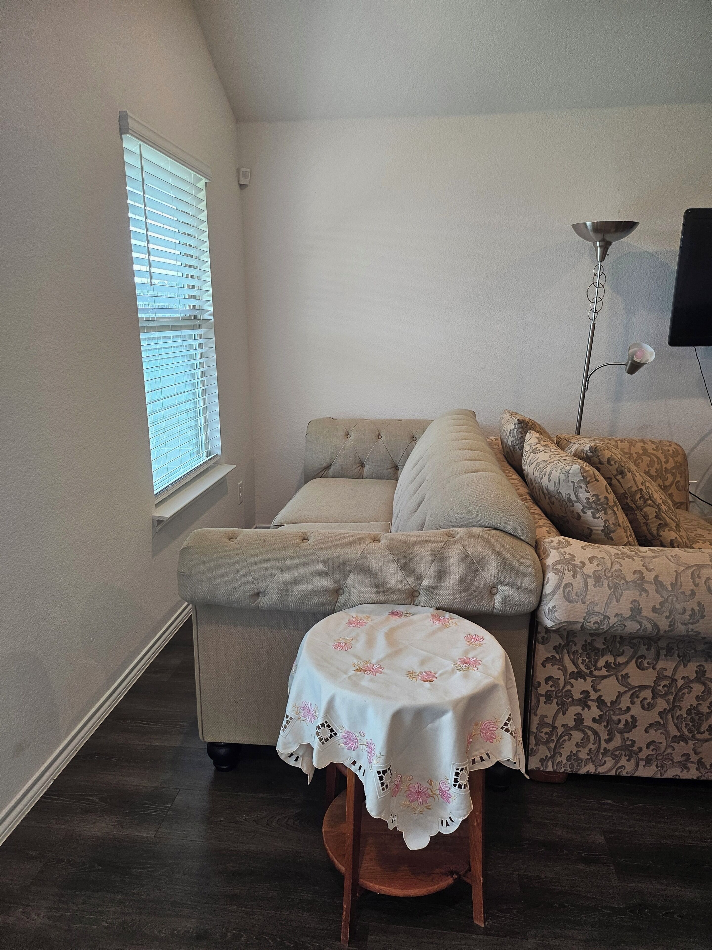 A cozy room with a sofa, a small round table with a floral tablecloth, a window with blinds, and a floor lamp.