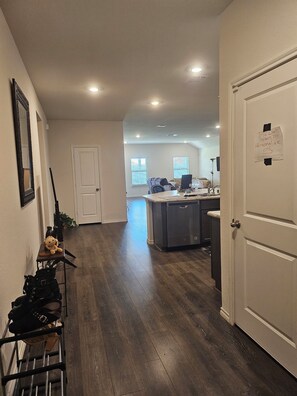 A hallway with a wooden floor, a white door, a small table with a teddy bear, and a rack with shoes.